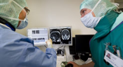 To grønnkledde leger med hetter og munnbinn studerer et bildeopptak av hjernen til en pasient, opptaket vises på en dataskjerm foran dem.