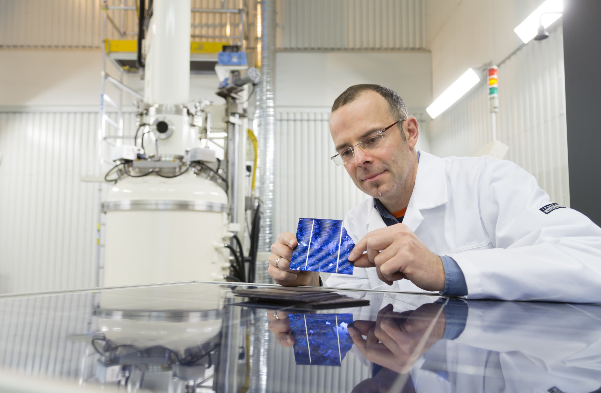 Mann i hvit labororiefrakk sitter i laboratorium mens han ser på en solcelle han holder i hendene