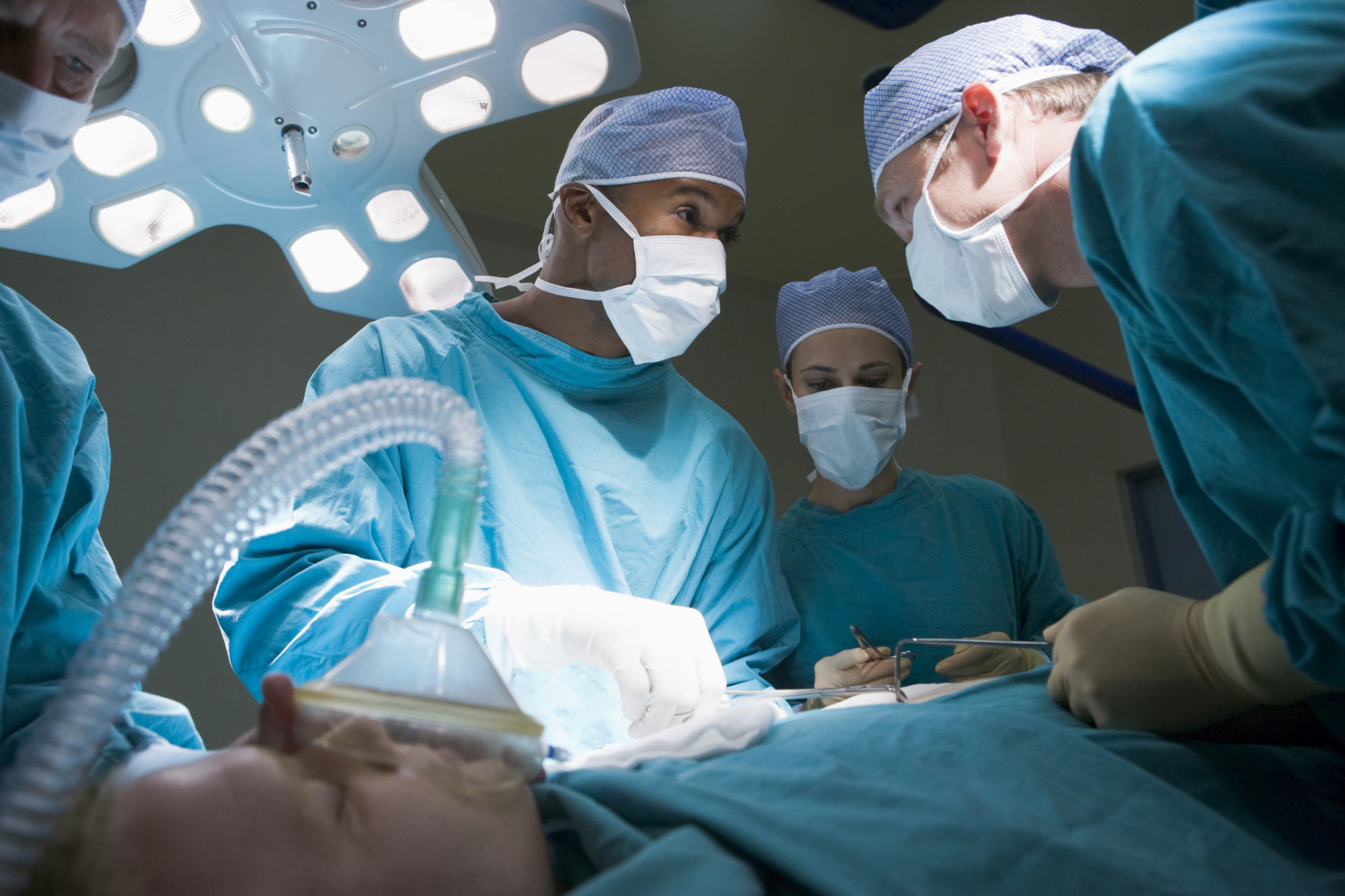 Kirurger med grønne klær og hvite munnbinn står bøyd over pasient ved operasjonsbord