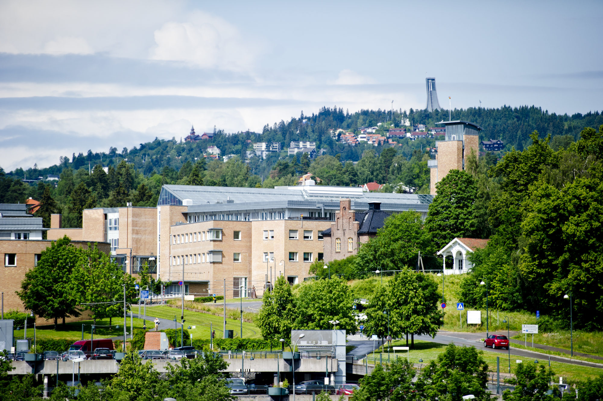 Bilde av lyserøde og beige sykehusbygg med Holmenkollåsen og tårnet på Holmenkollbakken i bakgrunnen.