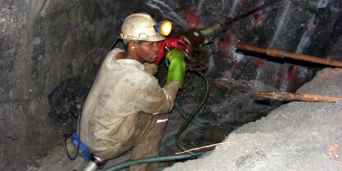 Gruvearbeider sittende på huk foran en bergvegg i gruve, med bormaskin foran seg