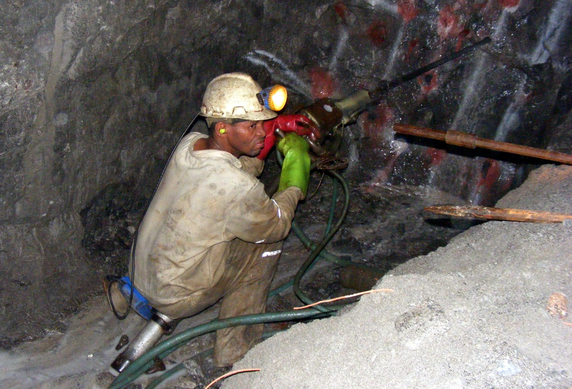 Gruvearbeider sittende på huk foran en bergvegg i gruve, med bormaskin foran seg
