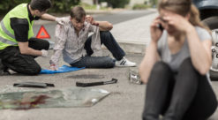 Ung jente og gutt sitter på asfalten med bilvrak i bakgrunnen og hjelpende voksenperson ikledd gul vest.