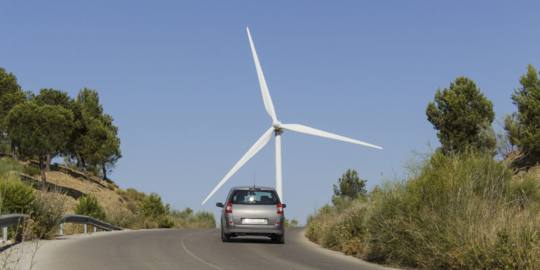 Personbil kjører mot bakketopp omgitt av gress og trær, og i horisonten ruver en vindmølle.