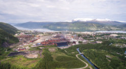 Flyfoto av Mo Industripark i Mo i Rana, grønne jorder i forgrunnen, fjord og fjell i bakgrunnen.