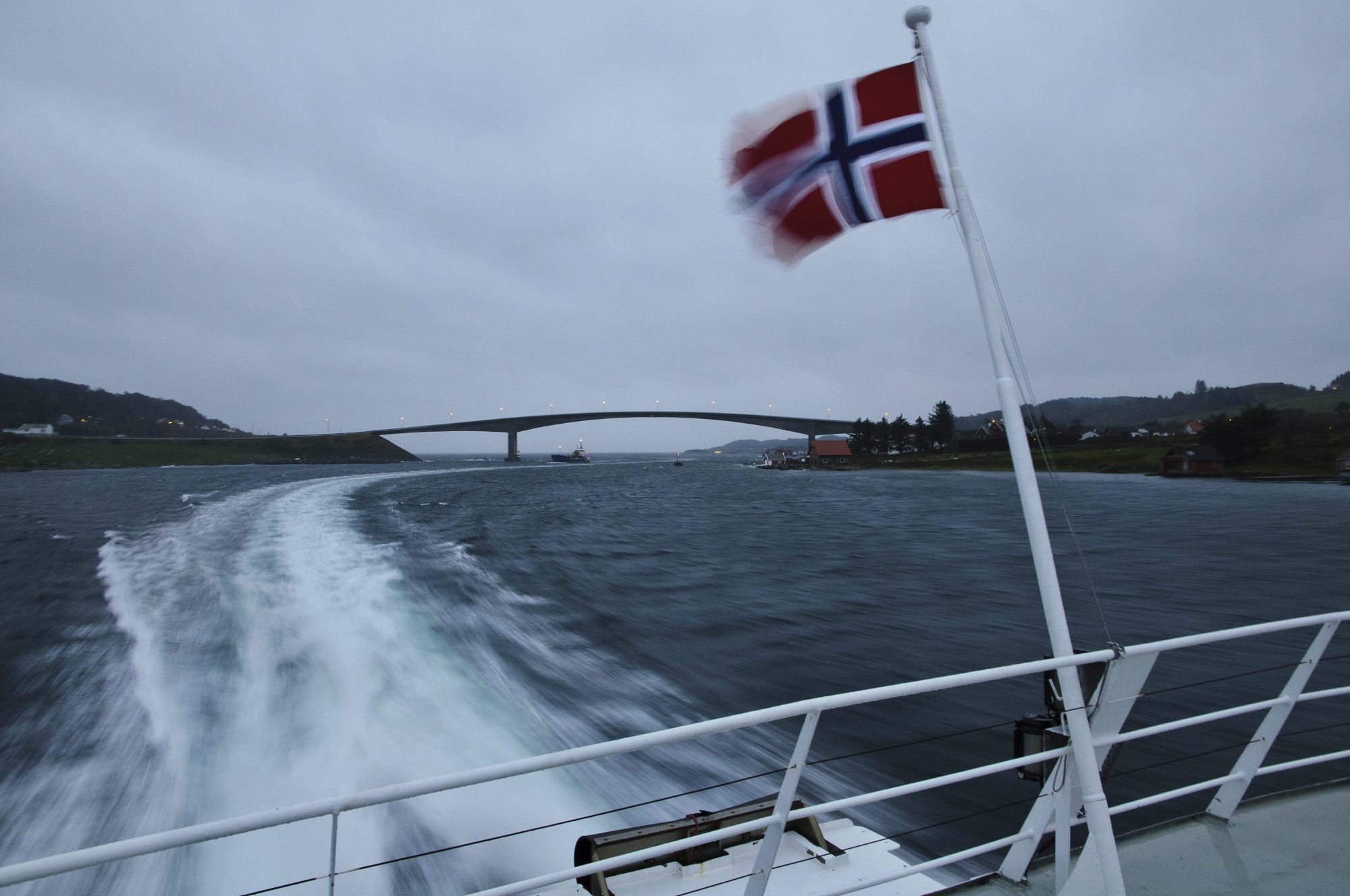 Bilde tatt fra bakdekket på en hurgibåt i overskyet vær. Det norske flagget vaier i forgrunnen. Kjølvann og kystlandskap vises i bakgrunnen.