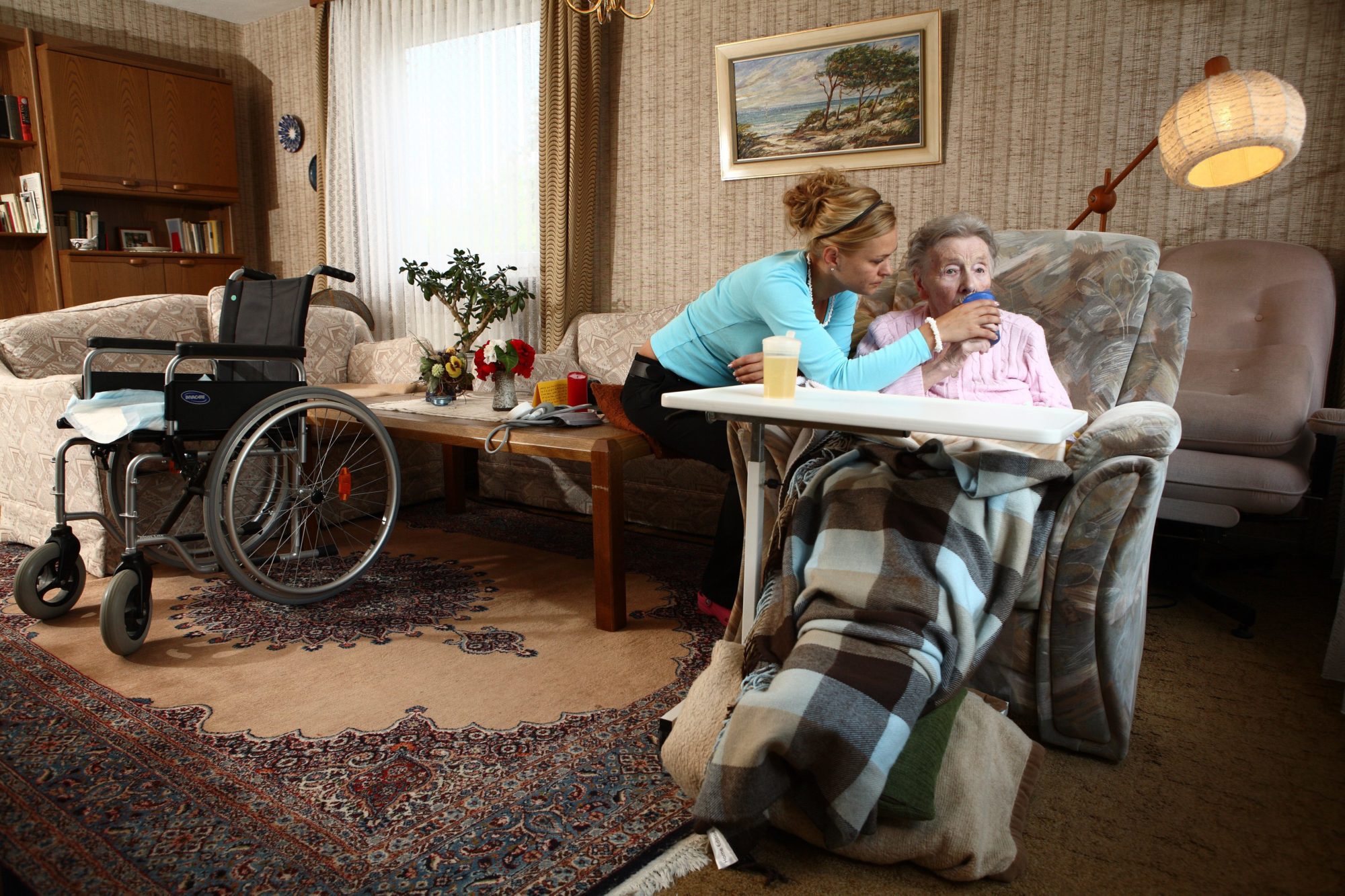 Kvinnelig hjemmesykepleier i turkis genser gir drikke til gammel kvinne i rullestolm i en stue med lysebrunt tapet og ditto gardiner.