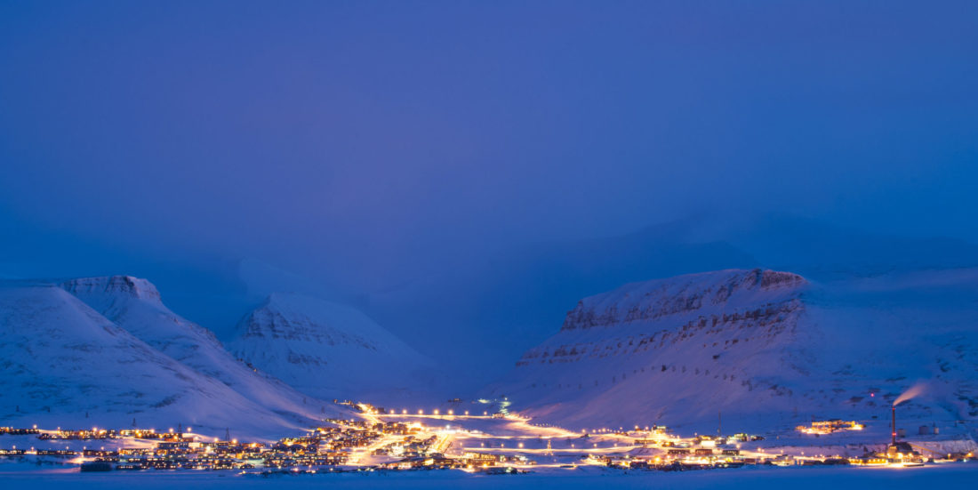 Nattbilde tatt fra fly over Longeyarbyen. Byen ses opplyst, med himmel og fjell i bakgrunnen, begge deler blå-lilla i kveldslyset.