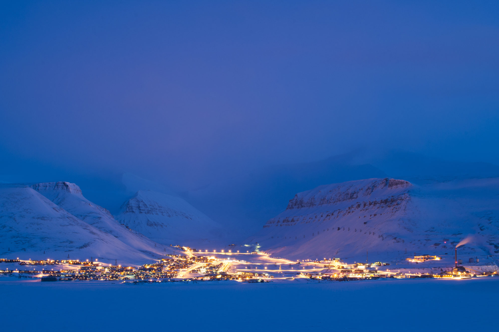 Nattbilde tatt fra fly over Longeyarbyen. Byen ses opplyst, med himmel og fjell i bakgrunnen, begge deler blå-lilla i kveldslyset.