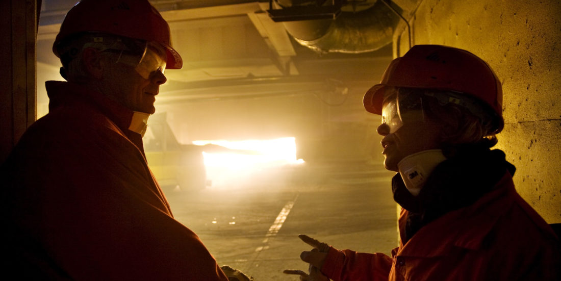 Silhuett av to mennesker med hjelm og kjeldress foran smelteovn der flammer ses gjennom åpen luke.