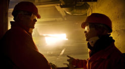 Silhuett av to mennesker med hjelm og kjeldress foran smelteovn der flammer ses gjennom åpen luke.