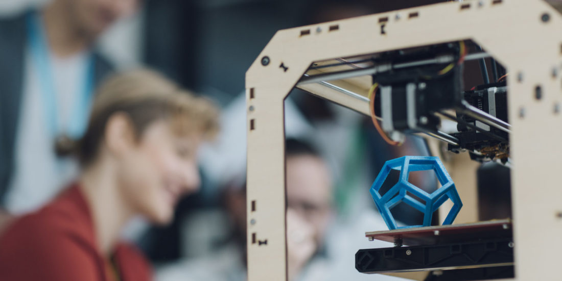 Blå metallkube printes ut av en 3Dprinter, mens mennesker i lab ser på.