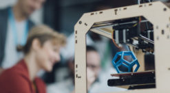 Blå metallkube printes ut av en 3Dprinter, mens mennesker i lab ser på.