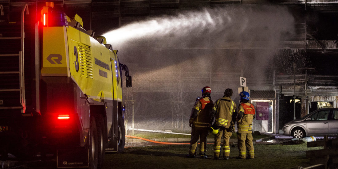 Gul lav flyplass-brannbil sprøyter vann på grått parkeringshus, tre brannmannskaper står ved siden av bilen.