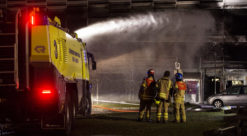 Gul lav flyplass-brannbil sprøyter vann på grått parkeringshus, tre brannmannskaper står ved siden av bilen.