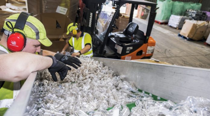 Industriarbeider med gulgrønn hjelm bøyer seg over container med resirkulerte glassklare plastflasker.