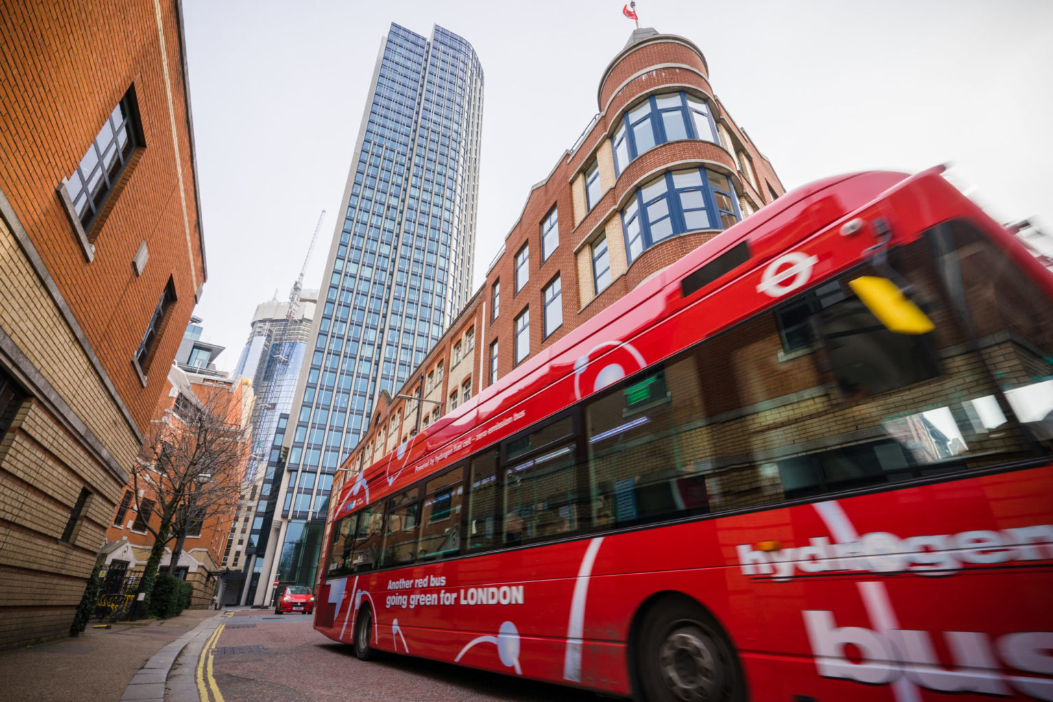 Rød dobbeltdekker buss med hvit påskrift "hydrogen bus" kjører gjennom London-gata med høyhus i bakgrunnen