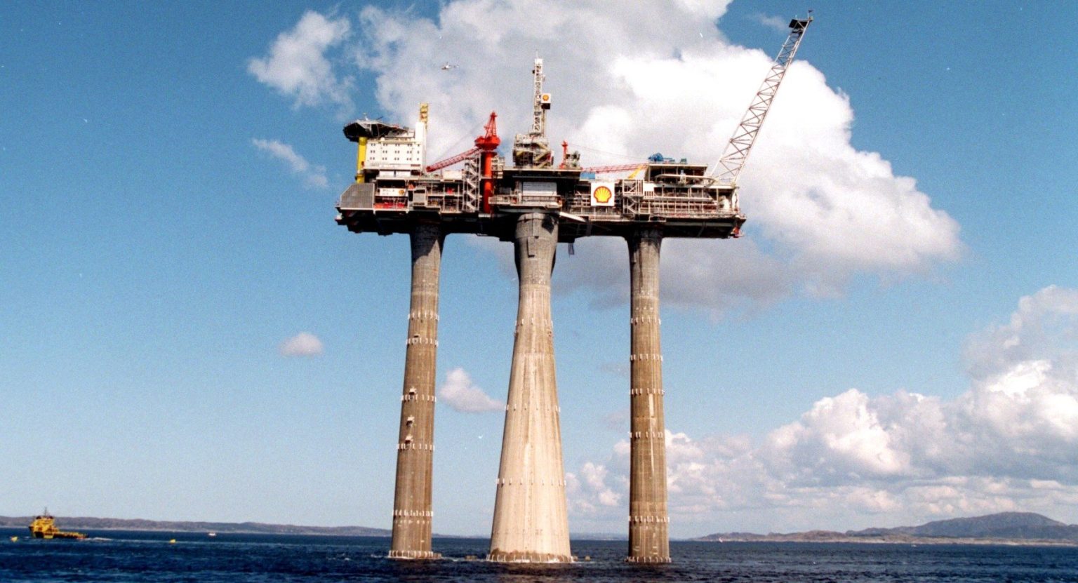 Høy betongplattform med tre lange bein slepes ut fra norskekysten under en blå himmel med stor hvit sky