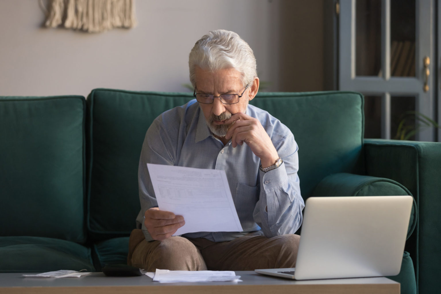 Eldre mann sitter i sofa med laptop på bordet foran seg mens han studerer er hvitt A4-ark.