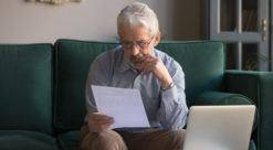 Eldre mann sitter i sofa med laptop på bordet foran seg mens han studerer er hvitt A4-ark.