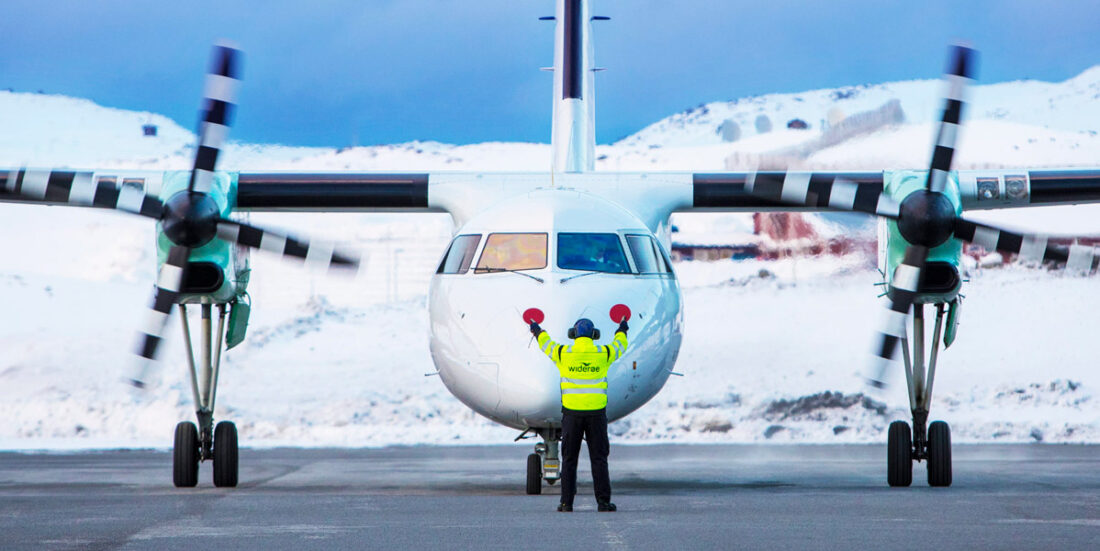 To-motors propellfly fra Widerøe fotografert etter landing i Hammerfest med vinterlandskap bak og gulkledt medlem av bakkemannskap foran flyet, med oransje spaker i hendene.