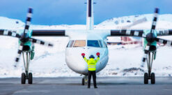To-motors propellfly fra Widerøe fotografert etter landing i Hammerfest med vinterlandskap bak og gulkledt medlem av bakkemannskap foran flyet, med oransje spaker i hendene.