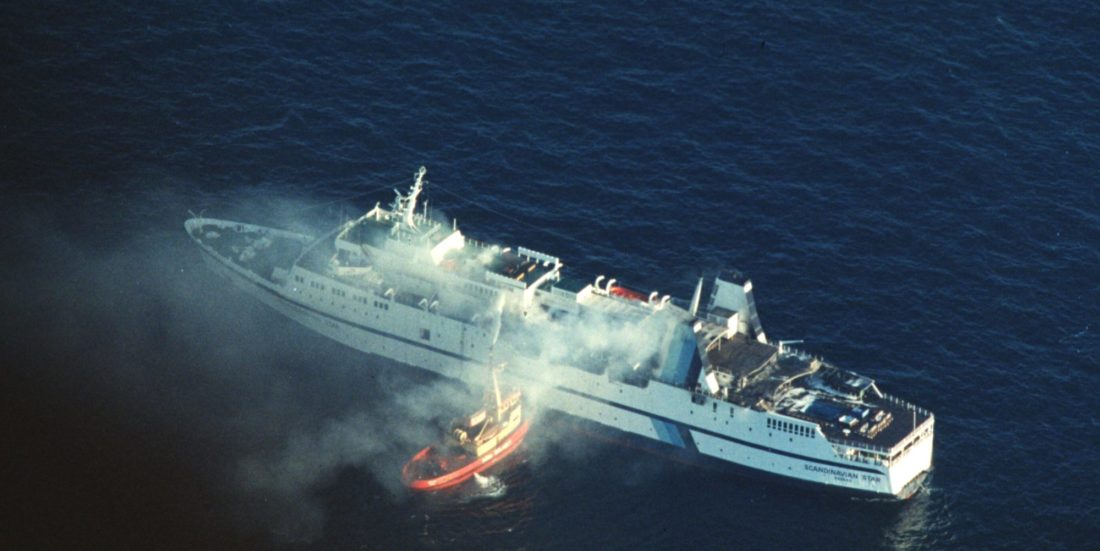 Luftfoto av brannen på Scandinavian Star, mindre sjøbrannsprøyte ved siden av det brennende skipet pøser på med vann.