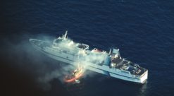 Luftfoto av brannen på Scandinavian Star, mindre sjøbrannsprøyte ved siden av det brennende skipet pøser på med vann.