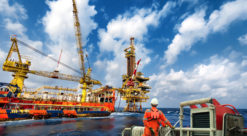 Offshore-arbeideer i oransje dress og hvit hjelm ses i forgrunnen, foran skip som utfører arbeid med en offshore-plattform.