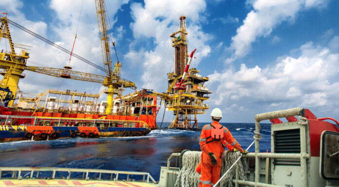 Offshore-arbeideer i oransje dress og hvit hjelm ses i forgrunnen, foran skip som utfører arbeid med en offshore-plattform.