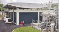Industrianlegg for vannelektrolyse / produksjon av grønt hydrogen fotografert på avstand med to medarbeidere i røde kjeldresser midt på bildet.