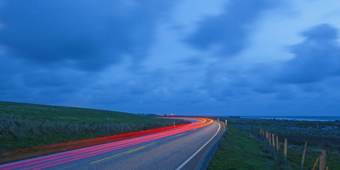 Lysstripe (lang lukkertid på kamera) langs riksvei nær kyst med mørkeblå himmel i bakgrunnen.