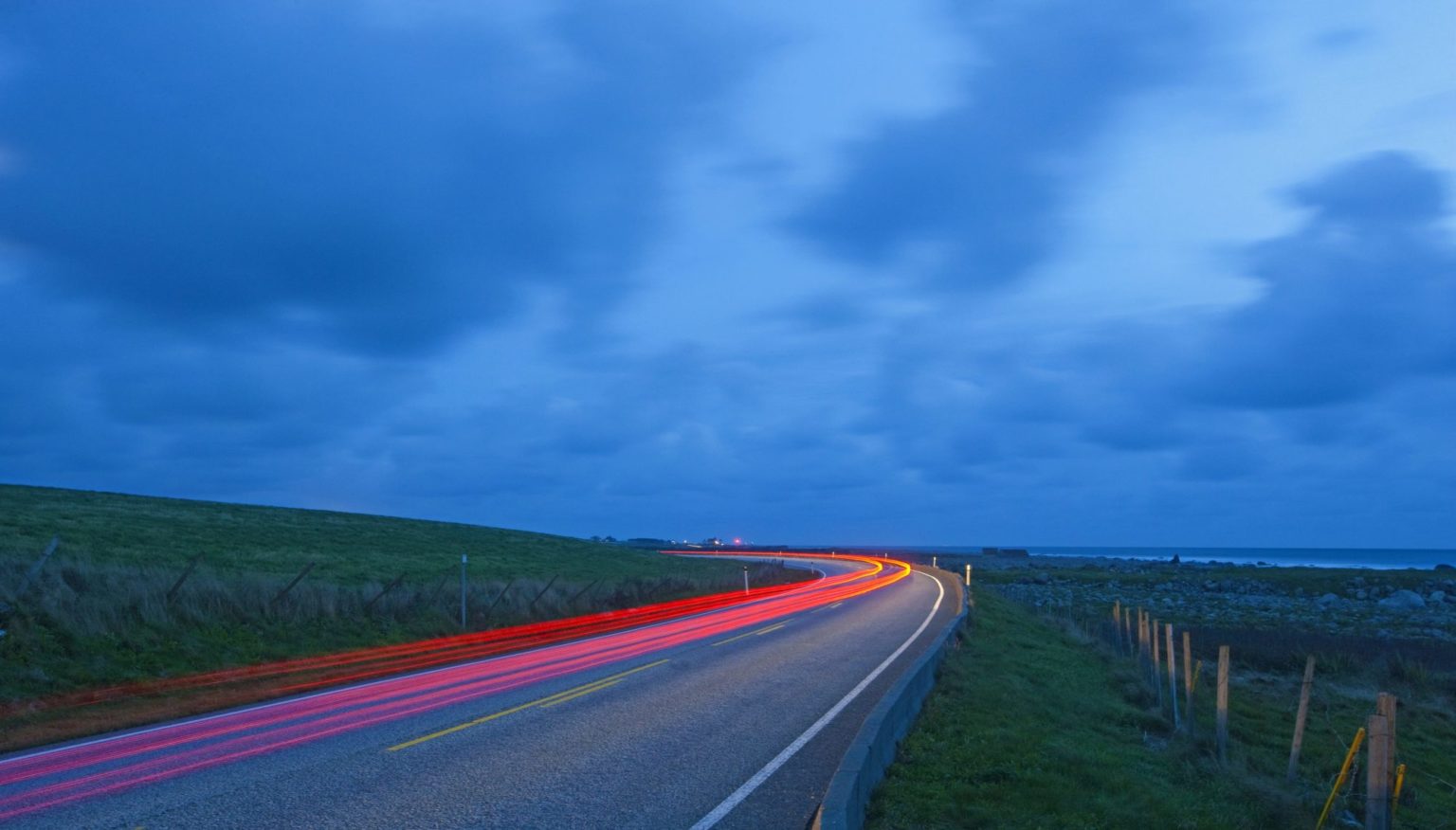 Lysstripe (lang lukkertid på kamera) langs riksvei nær kyst med mørkeblå himmel i bakgrunnen.