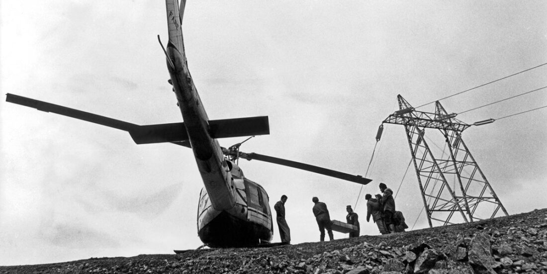 Svart-hvitt-foto som viser halen på helikopter som står på bakken foran kraftmaster og anleggsarbeidere.