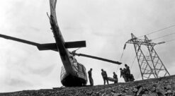 Svart-hvitt-foto som viser halen på helikopter som står på bakken foran kraftmaster og anleggsarbeidere.