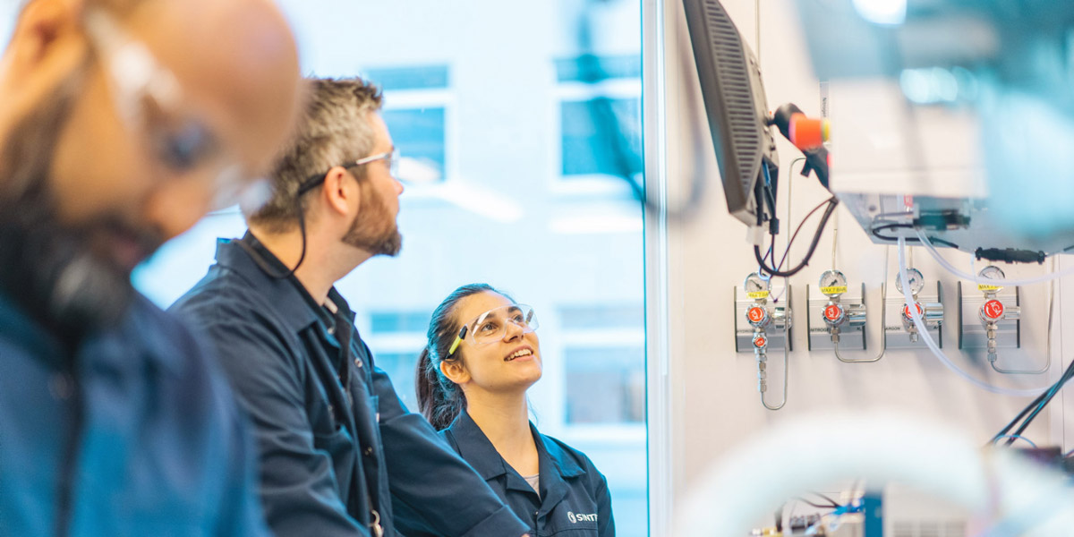 Tre forskere kledd i blå frakker og iført vernebriller ser opp på glassrør over laboratoriebenk.