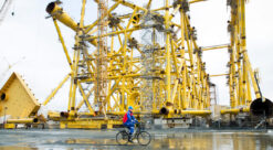 Svært gult stålunderstell til oljeplattform bygges på verftstomt. I forgrunnen: en mann på sykkel.