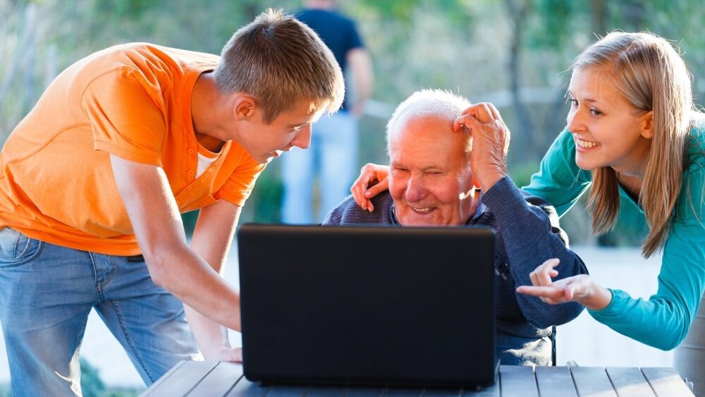 Hjernen. Bildet viser en eldre mann med en laptop. To unge mennesker viser noe på skjermen,.