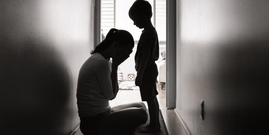 Svart hvitt foto der gråtende kvinne sitter foran et barn i en korridor. Begge ses i silhuett mot vindu i bakgrunnen.