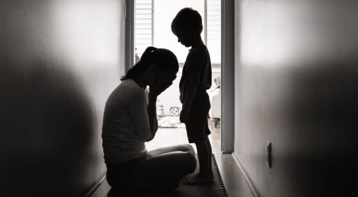 Svart hvitt foto der gråtende kvinne sitter foran et barn i en korridor. Begge ses i silhuett mot vindu i bakgrunnen.