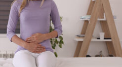 Bilde av kvinne i lys lilla genser og hvit bukse som sitter på venterom hos lege og holder hendene foran mageregionen.