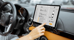 Bilde fra innsiden av bilkupe. En kvinnes hånd vises til høyre for rattet, der den trykker på et stort display med bilteknisk informasjon, blant annet om lading.