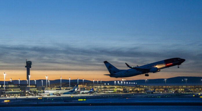 Fly tar av kveldsmørke. I bakgrunnen ses terminalbygning og tårn på Oslo Lutfhavn Gardermoen mot en oransje kveldshimmel