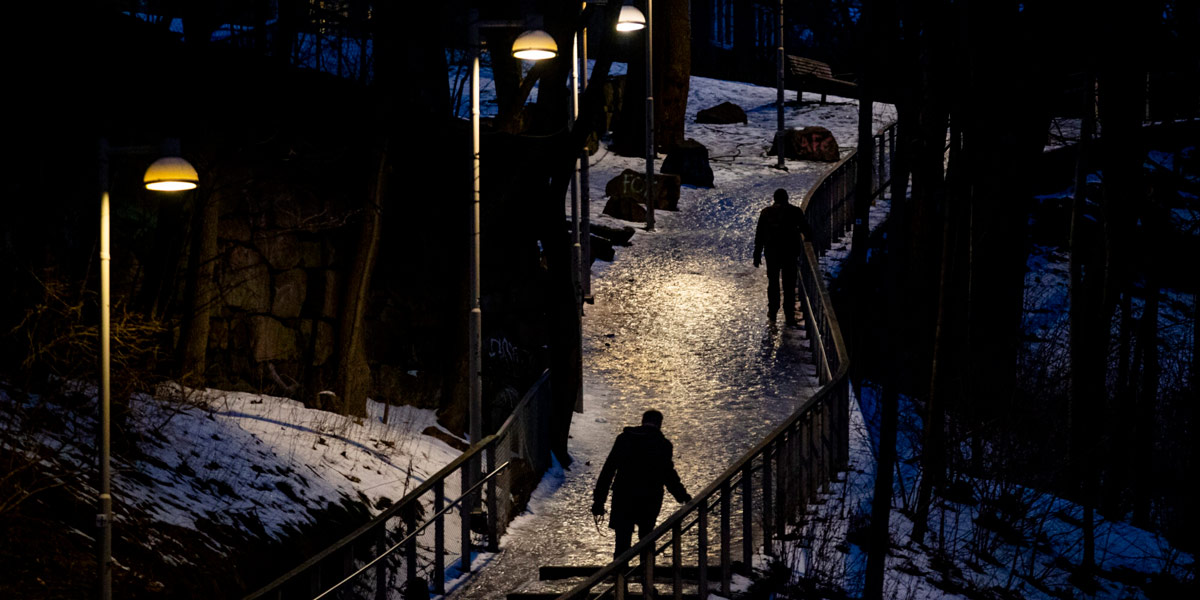 Kveldsbilde viser silhuetter av to mennesker som går på sti med hålke i skinnet fra gatelykter.