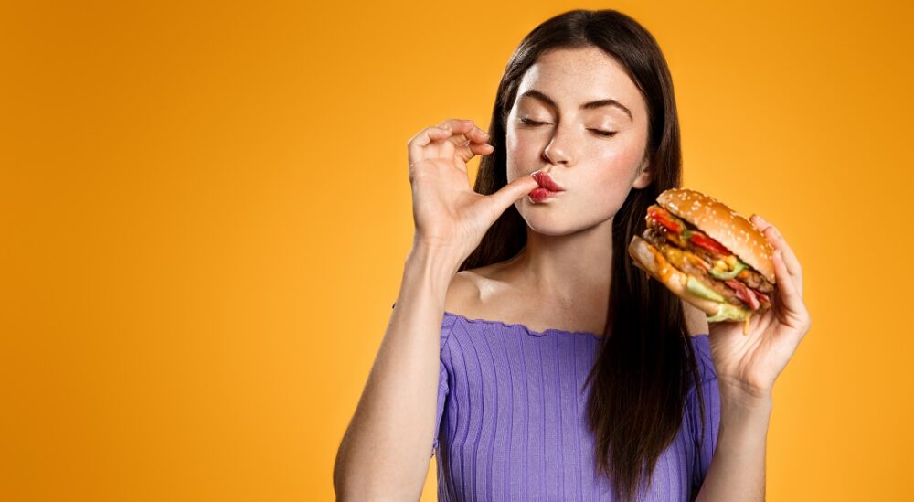 Kosthold. Bildet viser ei ung jente som slikker seg på fingerene og skal ha burger.