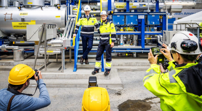 Bilde av to menn, i gule og blå kjeldresser, med hver sin hvite hjelm. De står i en fabrikkhall og er omgitt av fotograferer. Også disse bærer beskyttelseshjelmer.