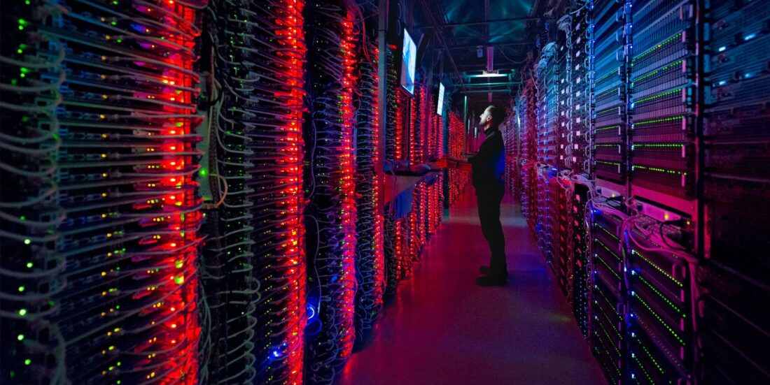 Bilde fra datasenter, mørkt rødskjær fra masse røde lyspunkter, mann ses i bakgrunnen