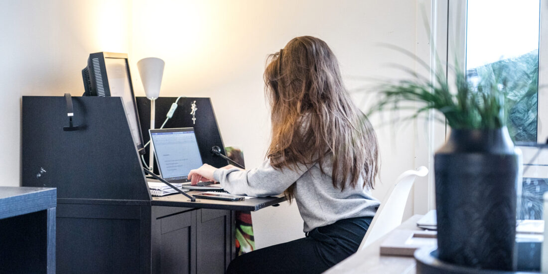 Ung dame med langt hår er fotografert fra siden/bakfra på arbeidsrom hjemme der hun sitter ved et skatoll og skriver på en pc. I forgrunnen til høyre ses en grønn plante i en krukke.