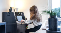 Ung dame med langt hår er fotografert fra siden/bakfra på arbeidsrom hjemme der hun sitter ved et skatoll og skriver på en pc. I forgrunnen til høyre ses en grønn plante i en krukke.