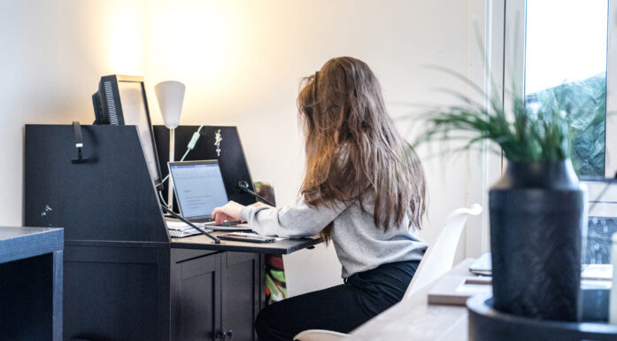 Ung dame med langt hår er fotografert fra siden/bakfra på arbeidsrom hjemme der hun sitter ved et skatoll og skriver på en pc. I forgrunnen til høyre ses en grønn plante i en krukke.